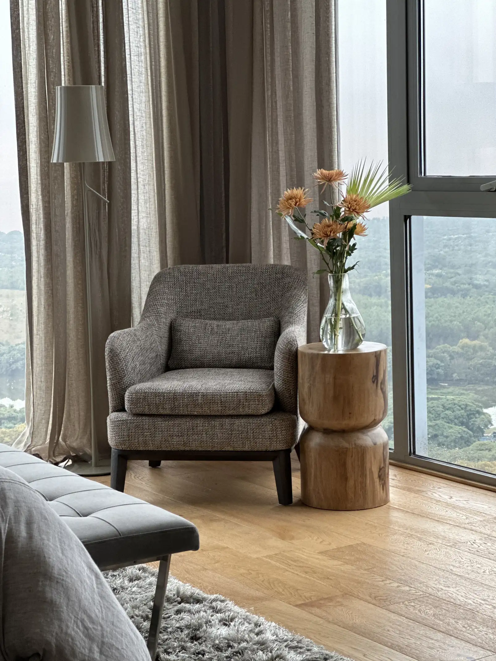 Rincón de lectura con sillón gris y flores neutras junto a ventana panorámica para proyectos residenciales que priorizan bienestar y calma.