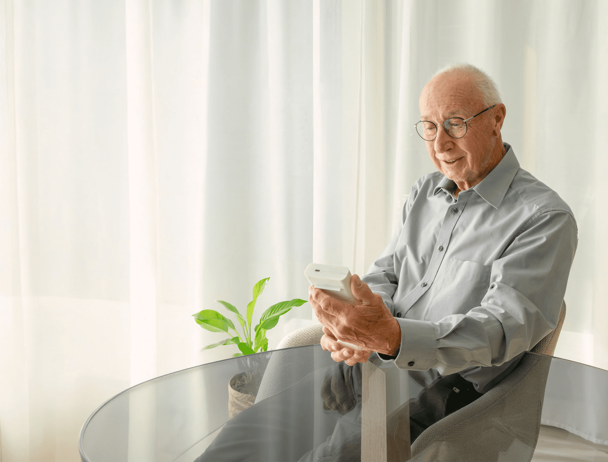 A smiling elderly man, wearing glasses, sitting at a table and holding a white smart medication dispenser (Medicase) in his hands.