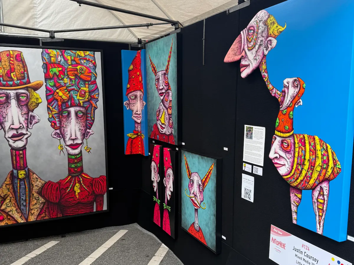 Colorful surreal mixed-media artwork displayed inside an artist booth at Artisphere in downtown Greenville, featuring abstract human figures with exaggerated faces and vivid neon colors.