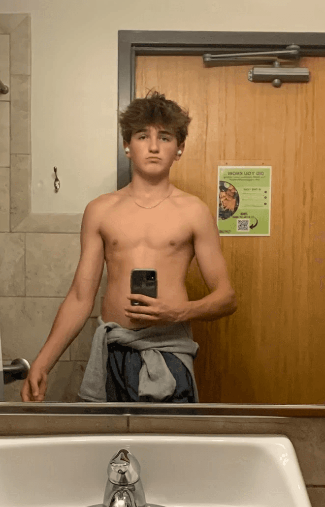 A man with short hair and a beard takes a mirror selfie in a dimly lit bathroom, wearing a green t-shirt and holding a smartphone.