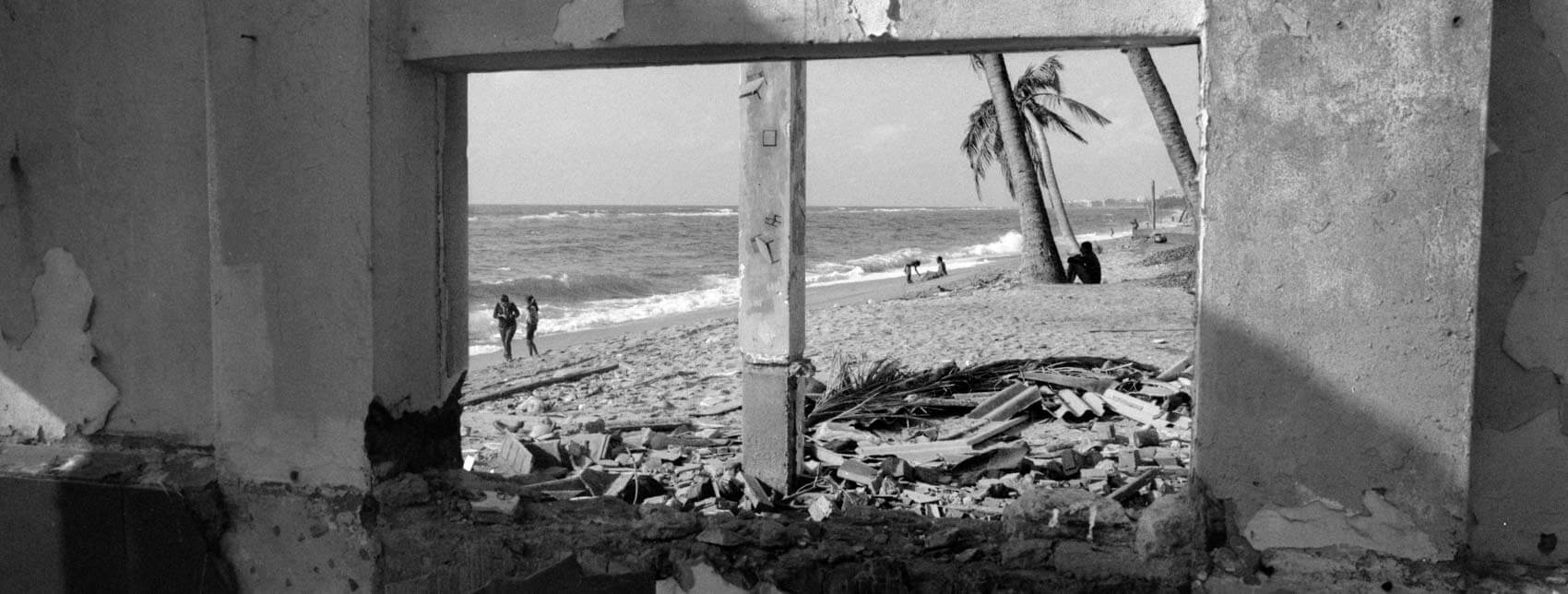 A Black and white film photograph of people walking along a soft Colombo beach, seen through the rough opening of an abandoned concrete house in Sri Lanka. Frame within a frame composition shot on Ilford Delta 400 with a Hasselblad XPan and 45mm lens.