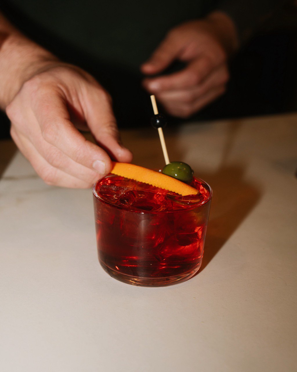 A bartender garnishes a vibrant red cocktail with an orange slice, green olive, and black cherry on a toothpick, emphasizing a classic and sophisticated presentation in a dimly-lit setting.