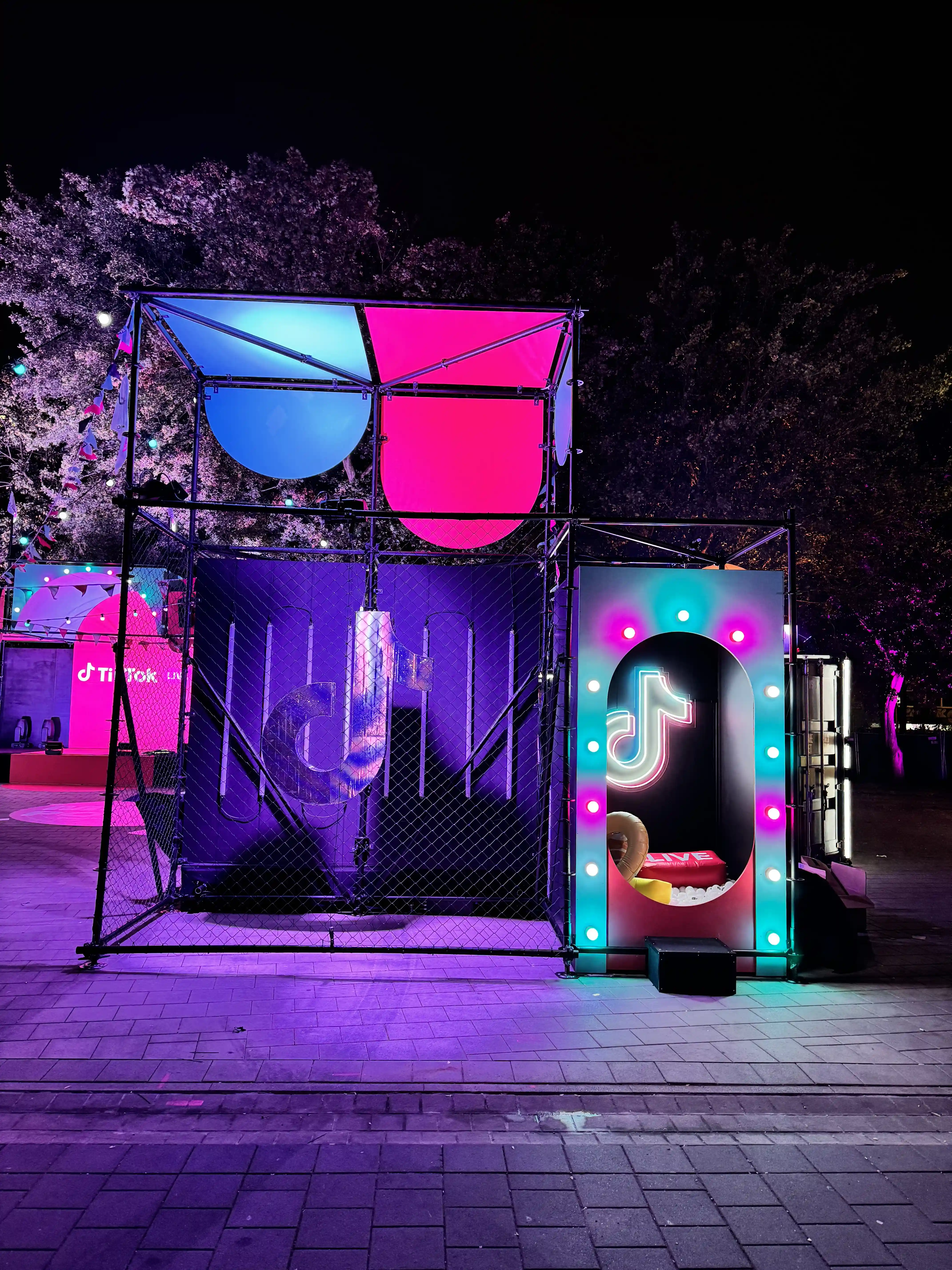 Night view of the TikTok scaffold installation with glowing arch photo booth and pink and blue canopies.