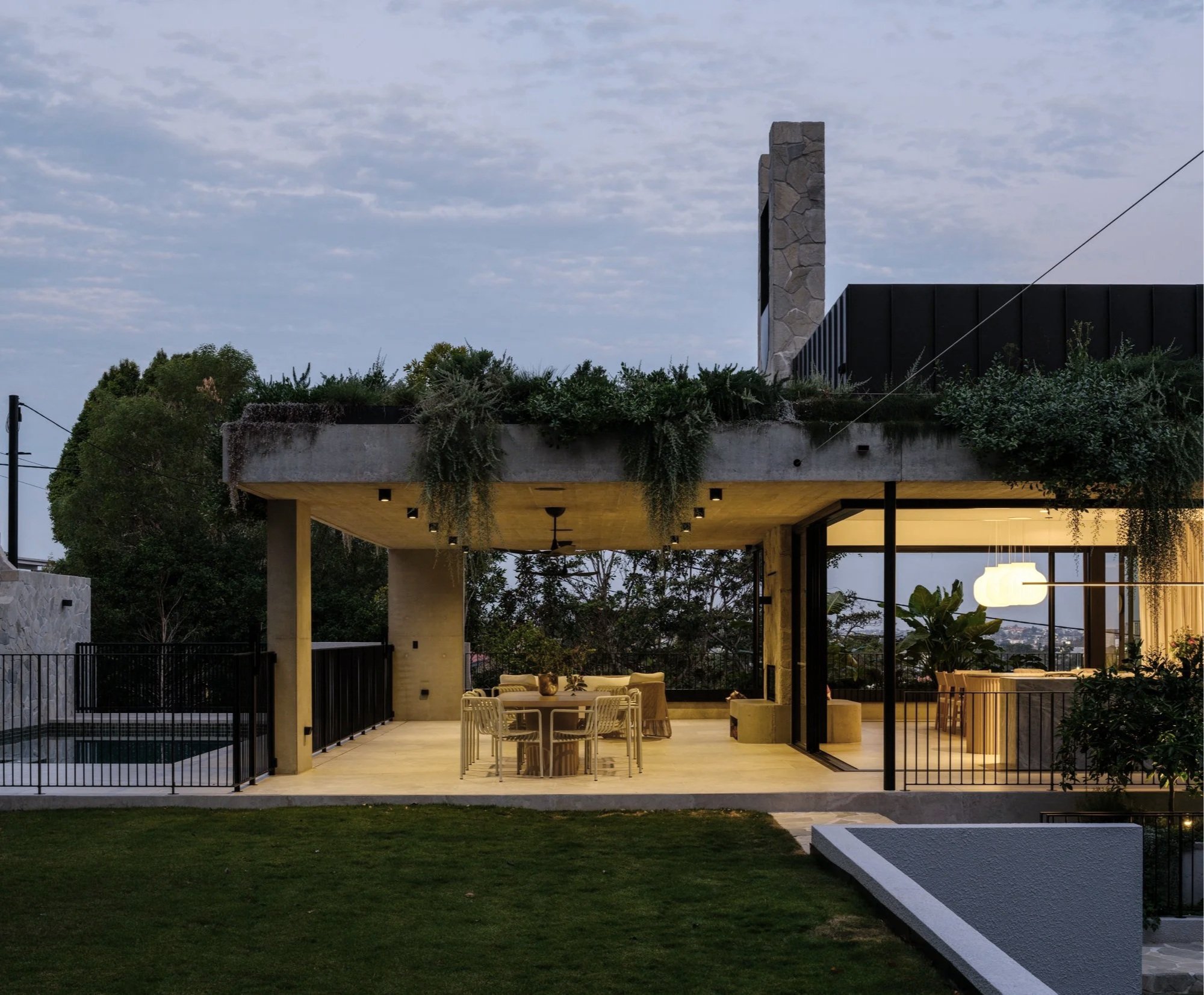 Covered outdoor terrace with concrete structure, integrated greenery, and open connection to indoor dining at dusk.