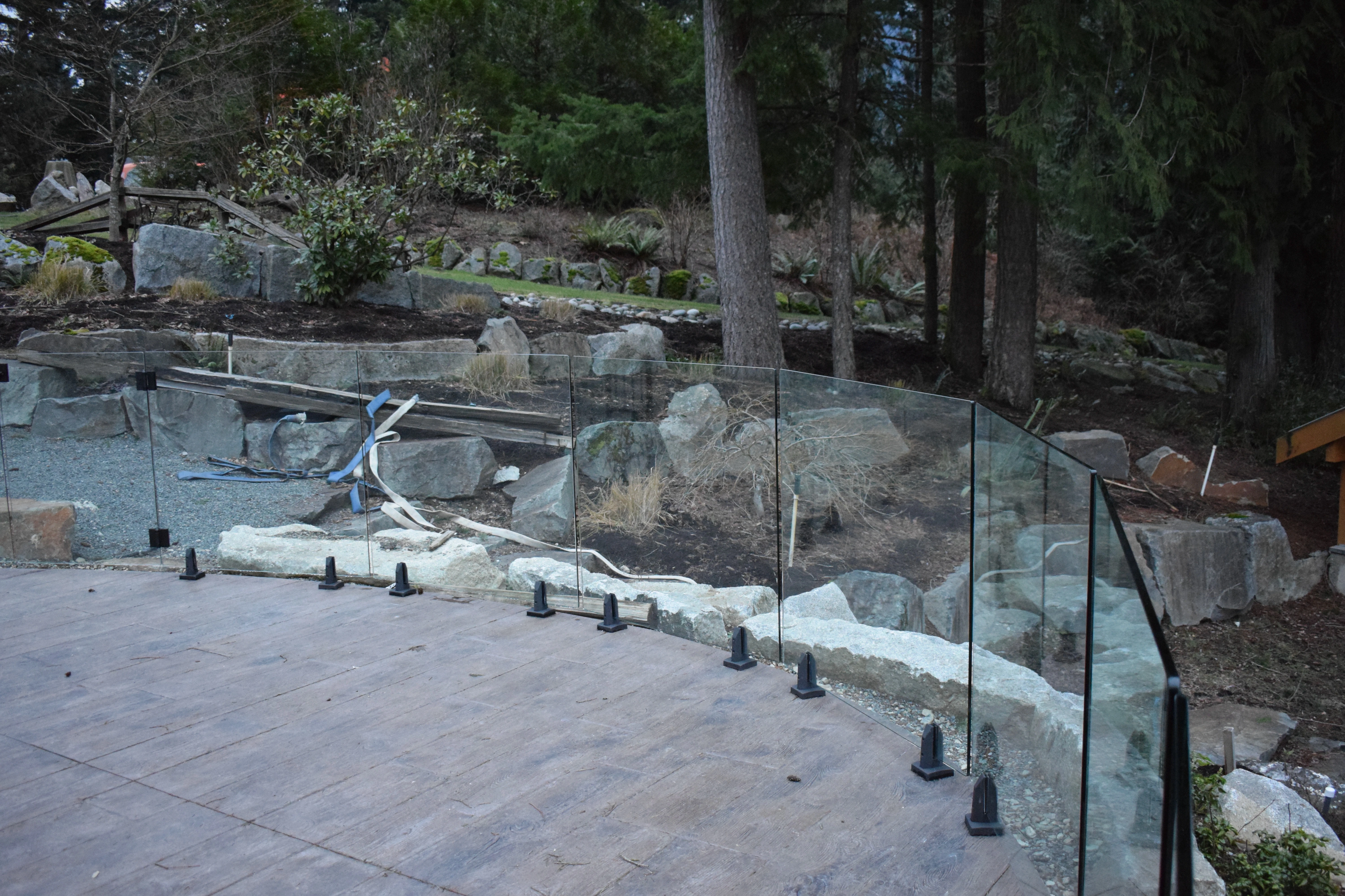 A curved run of frameless glass panels enclosing a stamped concrete patio, each panel secured at the base with individual black spigot clamps bolted along the paved edge. Large granite boulders and tall evergreen trees rise behind the railing, with construction materials still visible on the ground, suggesting a recently completed installation in a wooded BC backyard.