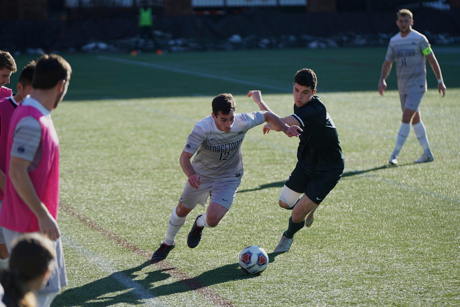 A soccer player in a grey "Georgetown" jersey dribbles the ball while a defender in a dark green kit attempts to tackle.