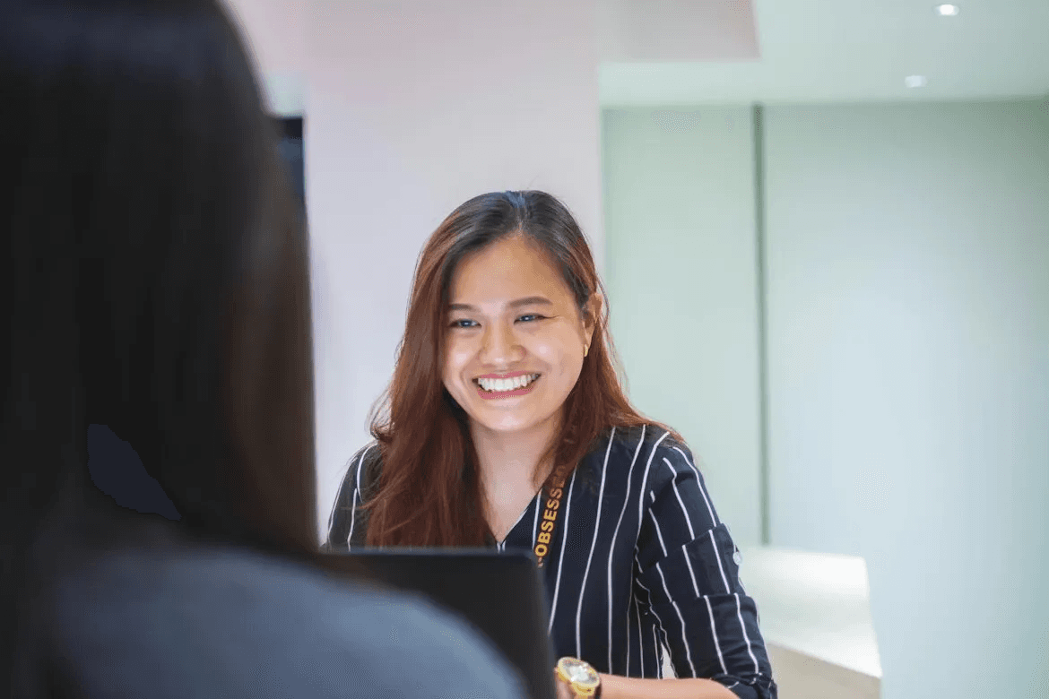 A woman with long auburn hair smiling warmly across a table during a meeting or interview, with another person visible in the foreground