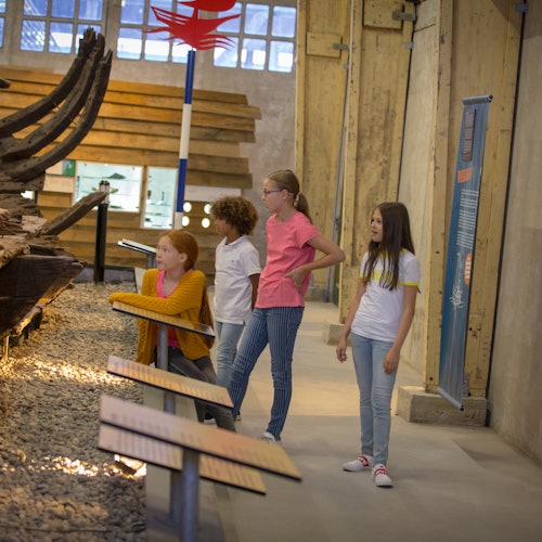 四个孩子站在博物馆里，观看一个古老木船展品，四周有信息面板。