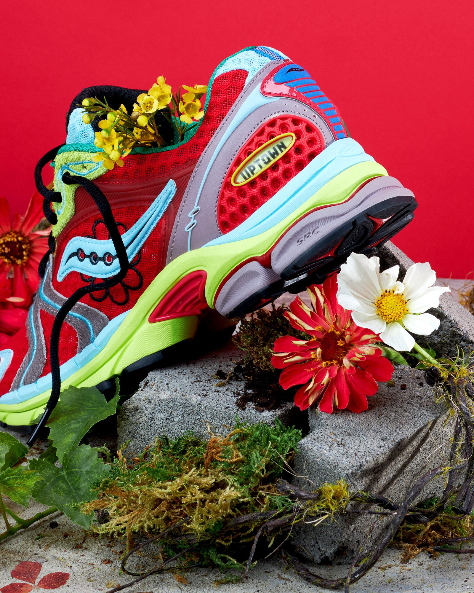 Close-up of a red Jae Tips x Saucony sneaker midsole and heel, framed with flowers and brick texture.