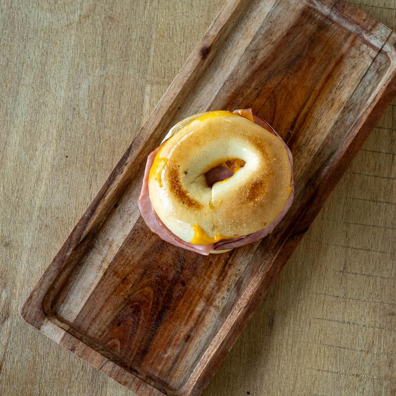 A rustic outdoor table setting featuring a charcuterie arrangement of cheese and sliced salami alongside a large bowl of uncooked pasta, a bottle of olive oil, and a glass of white wine, all against a dark, vine-covered background.