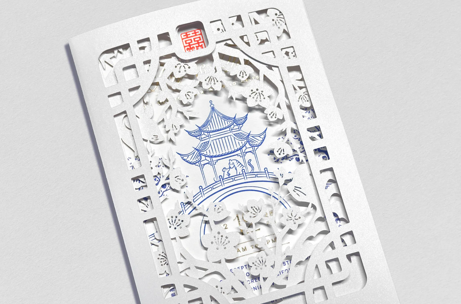 Closed tri-fold wedding brochure featuring decorative, laser cut side panels folding over and framing the center invitation card with chinese-porcelain-inspired illustration of a pagoda with a rat and tiger