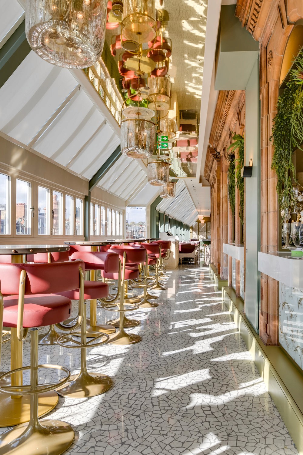 A vibrant bar and dining area with red barstools, patterned flooring, and glass ceilings adorned with chandeliers and greenery.