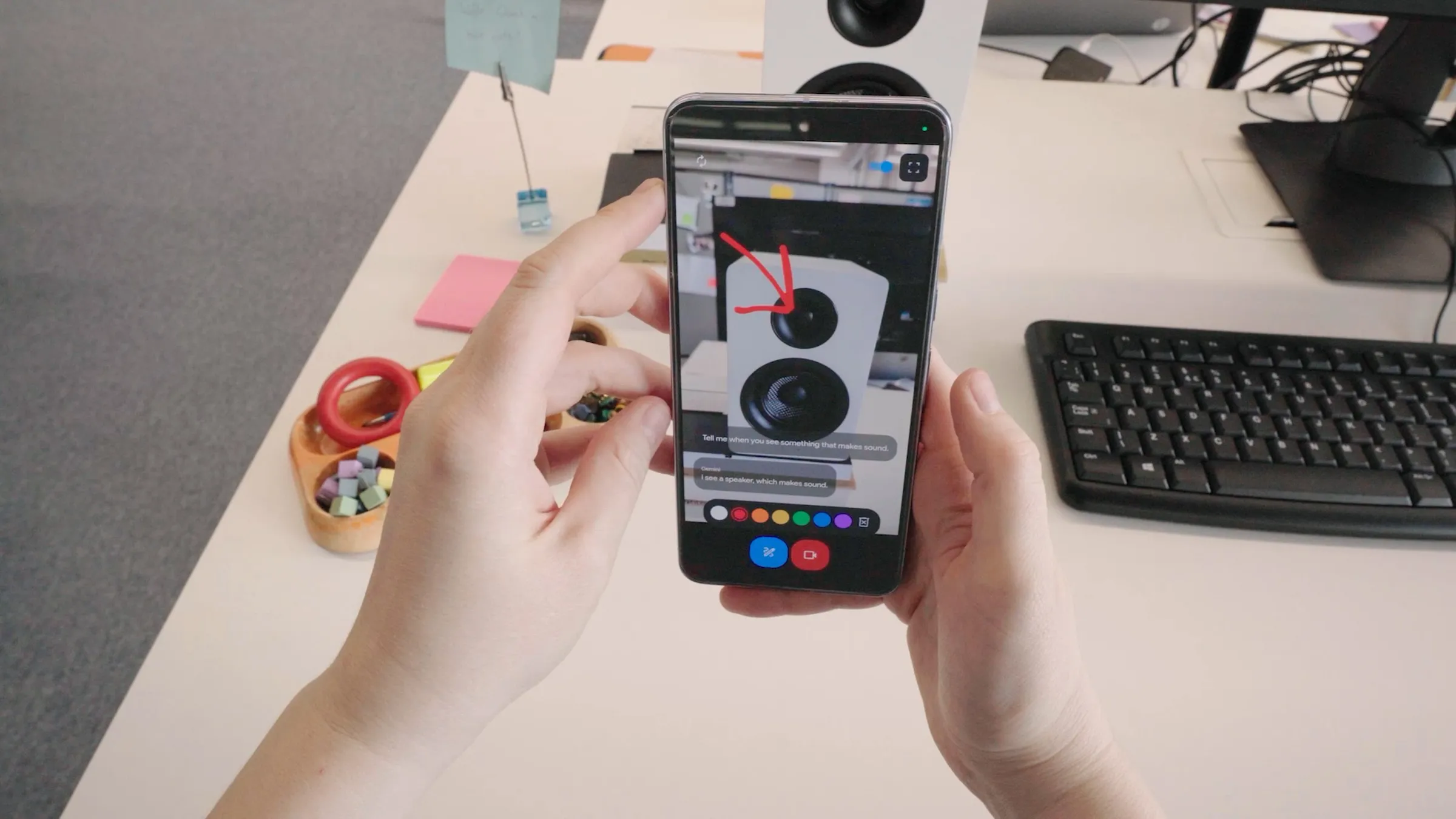 Person holding a smartphone displaying an image of a speaker with a red arrow pointing to it. Various objects, including a keyboard, sticky notes, and a bowl of small items, are on the desk. The scene suggests integration with universal digital assistants like Google Gemini Pro.