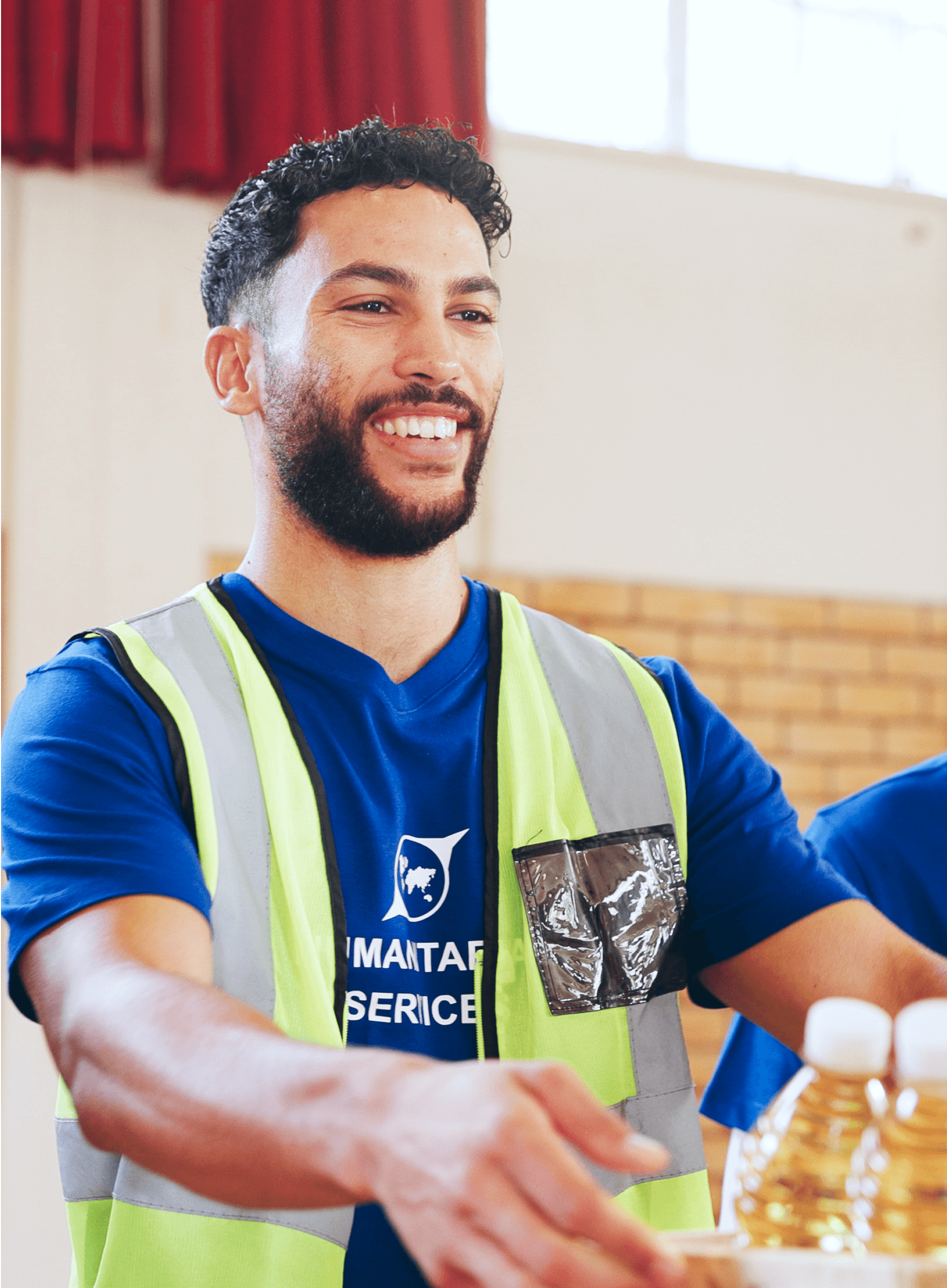 A smiling man in a blue shirt and yellow vest holds a stack of items, engaged in an activity indoors.