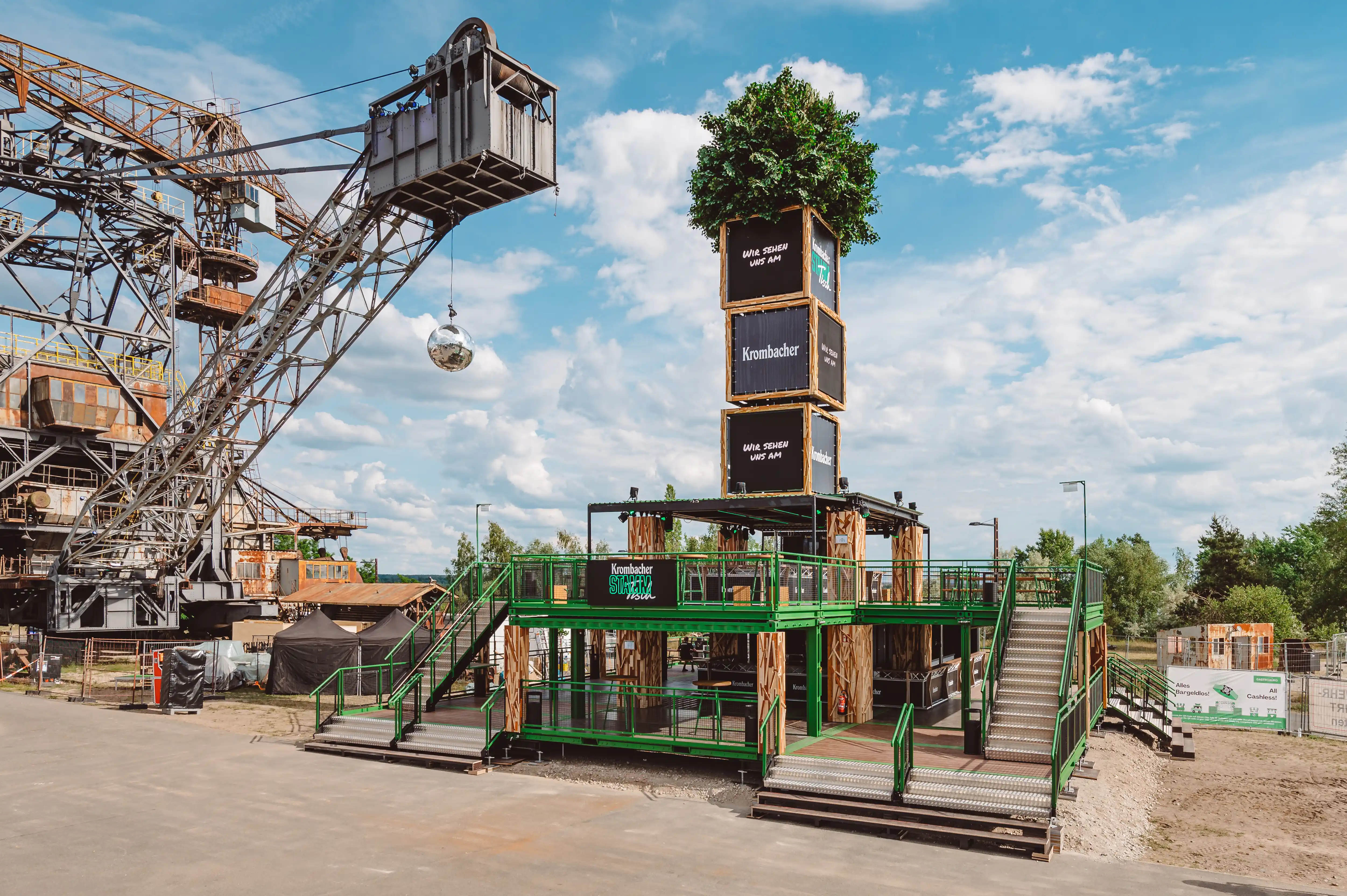 Two-storey KROMBACHER festival structure with green steel railings, wooden stairs and a crate tower crowned by a living tree, next to a giant bucket excavator.