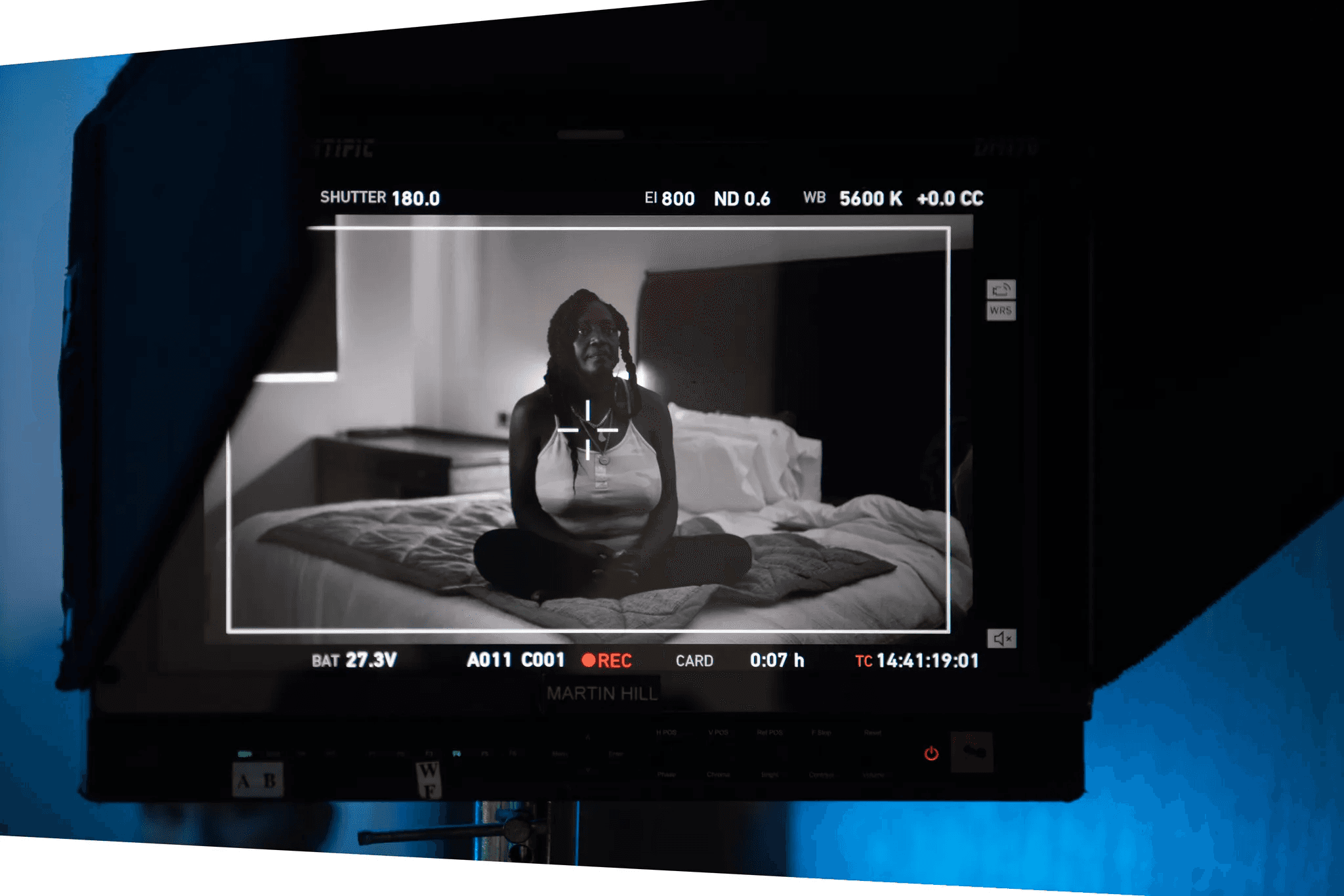 A video camera screen showing a black-and-white view of a woman sitting cross-legged on a bed during a recorded interview or testimonial, with studio lighting and equipment visible around the monitor.