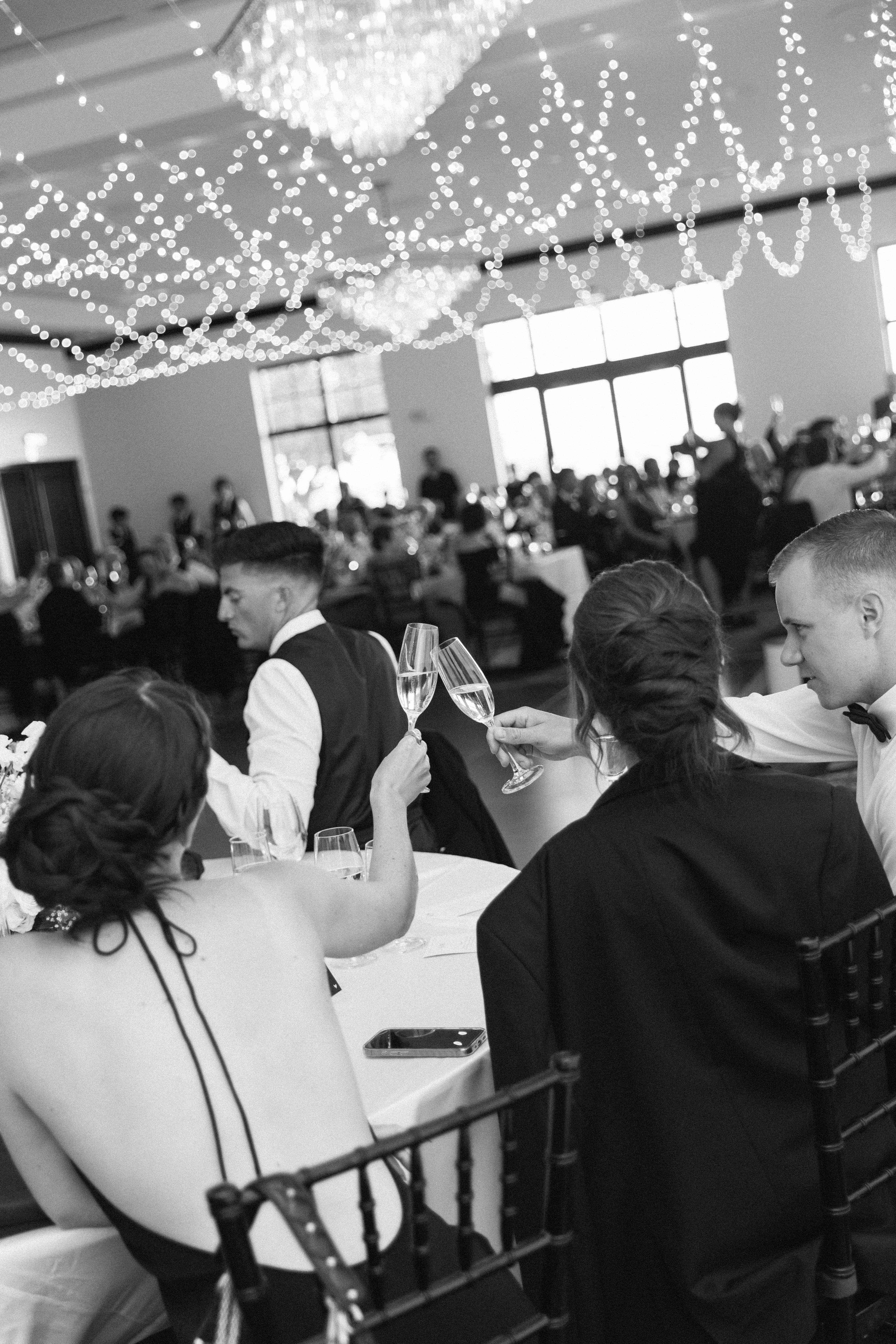 A black-and-white photo shows a group of people at a table, raising glasses in a toast. String lights hang from the ceiling in the background.