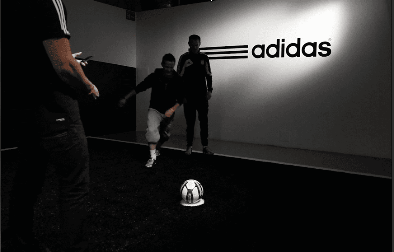 Three people in a dimly lit room engage with a stationary soccer ball on a black floor, beneath an illuminated "Adidas" sign on the wall; one person is in mid-action, seemingly preparing to kick the ball.