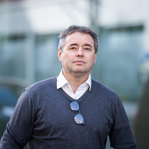 A man in a dark sweater stands outdoors, looking confidently at the camera against a blurred background.