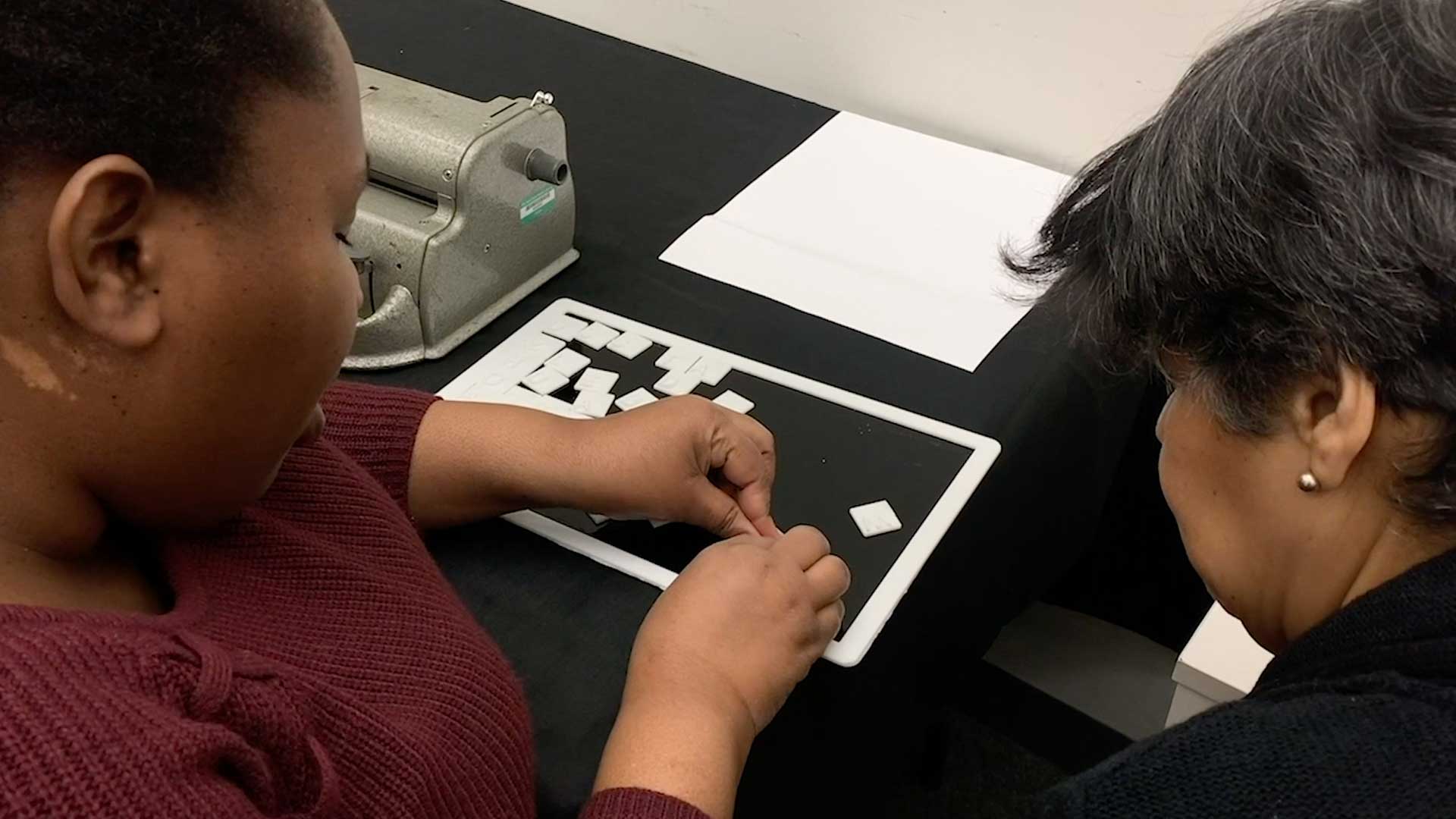 Two people are seated at a black table. The person on the left wears a maroon sweater and touches a white tablet displaying a grid of squares. The person on the right wears a black top and has dark hair. A silver toaster and a white sheet of paper are on the table between them. The background is a plain white wall.
