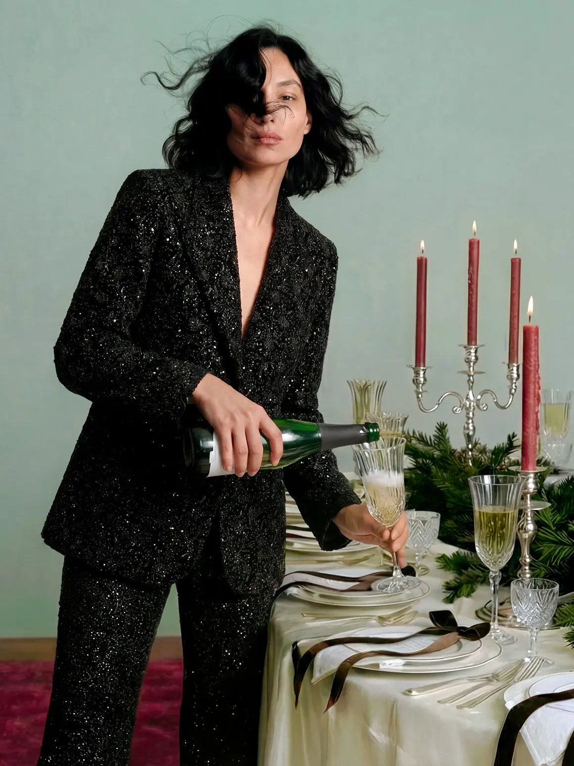 A person in a shimmering black suit pours champagne into a crystal flute on a lavishly set dining table adorned with red candles and elegant greenery, embodying a high-end fashion brand atmosphere.