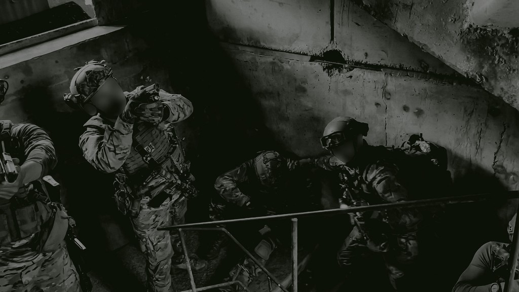 Soldier in camouflage uniform and helmet kneeling in a sandy area, aiming a rifle.