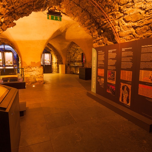A stone museum exhibit room with informational panels, an archway, glass display cases, and a green exit sign above.