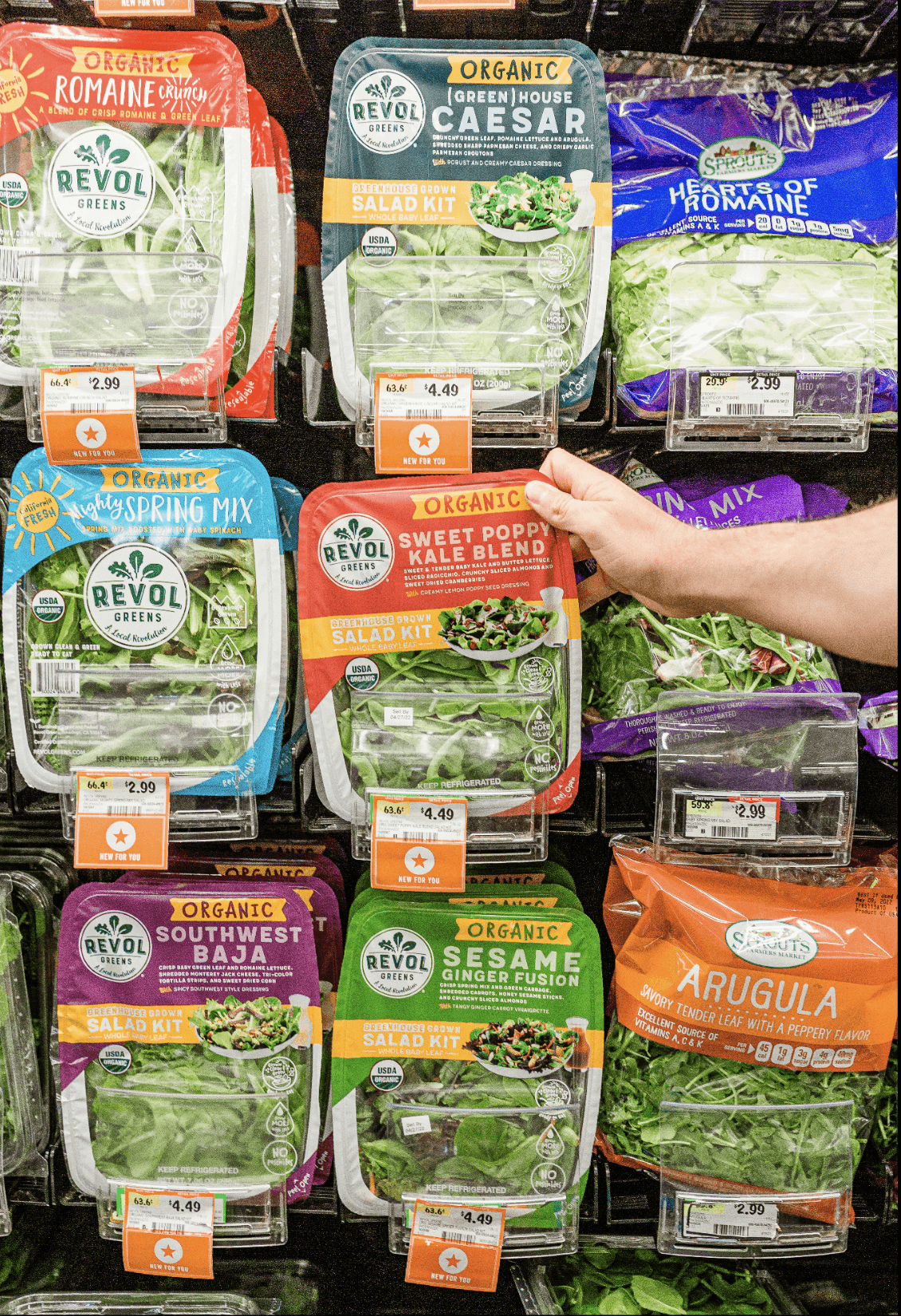 Refrigerated grocery shelf stocked with Revol Greens organic salad kits and greens, with a shopper reaching for the Sweet Poppy Kale Blend.