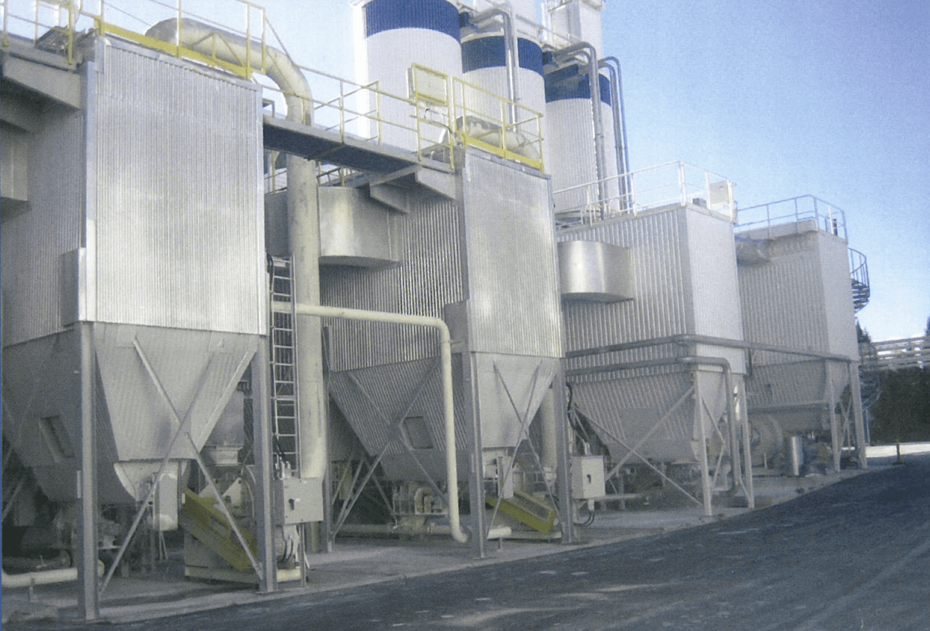 Industrial storage silos lined up in a facility against a clear blue sky, showcasing metal structures and pipes.