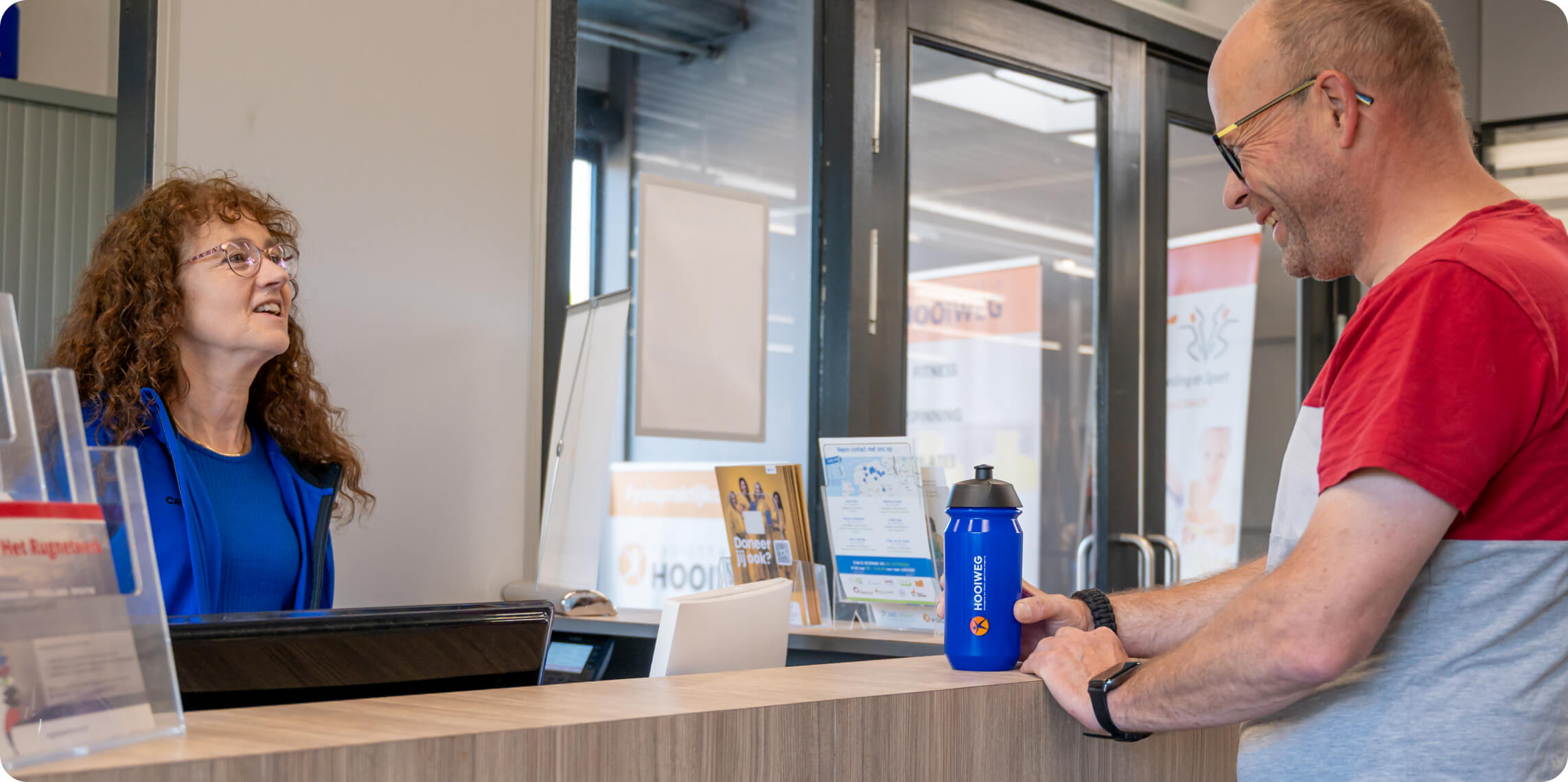 A woman with curly hair smiles at a man in a red and gray shirt holding a blue water bottle, as they interact at a modern reception desk with brochures and signs.