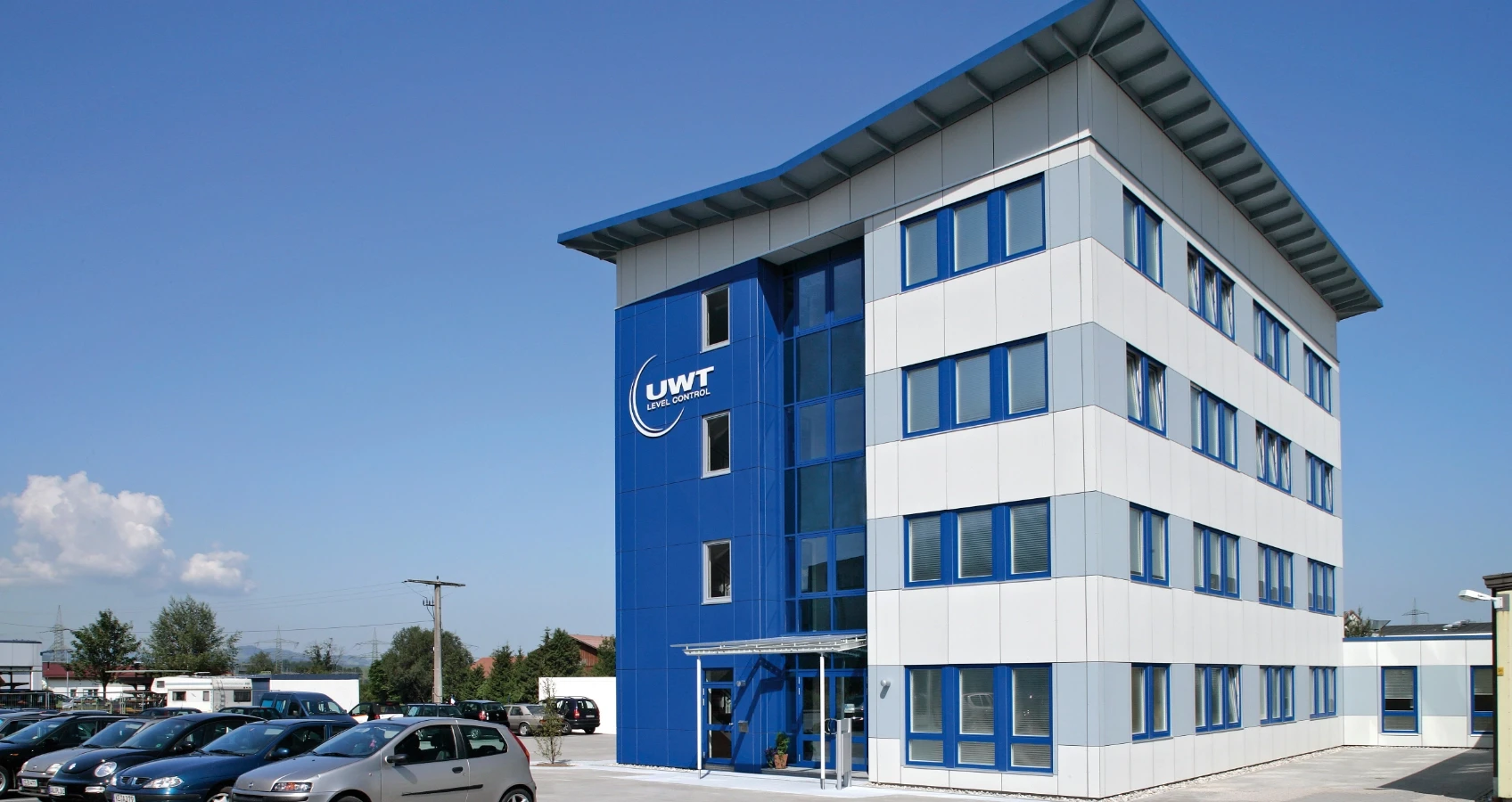 Modernes Bürogebäude mit blauen und weißen Fassadenpaneelen, blauen Fensterrahmen und dem Firmenlogo "UWT LEVEL CONTROL" auf der blauen Fläche.