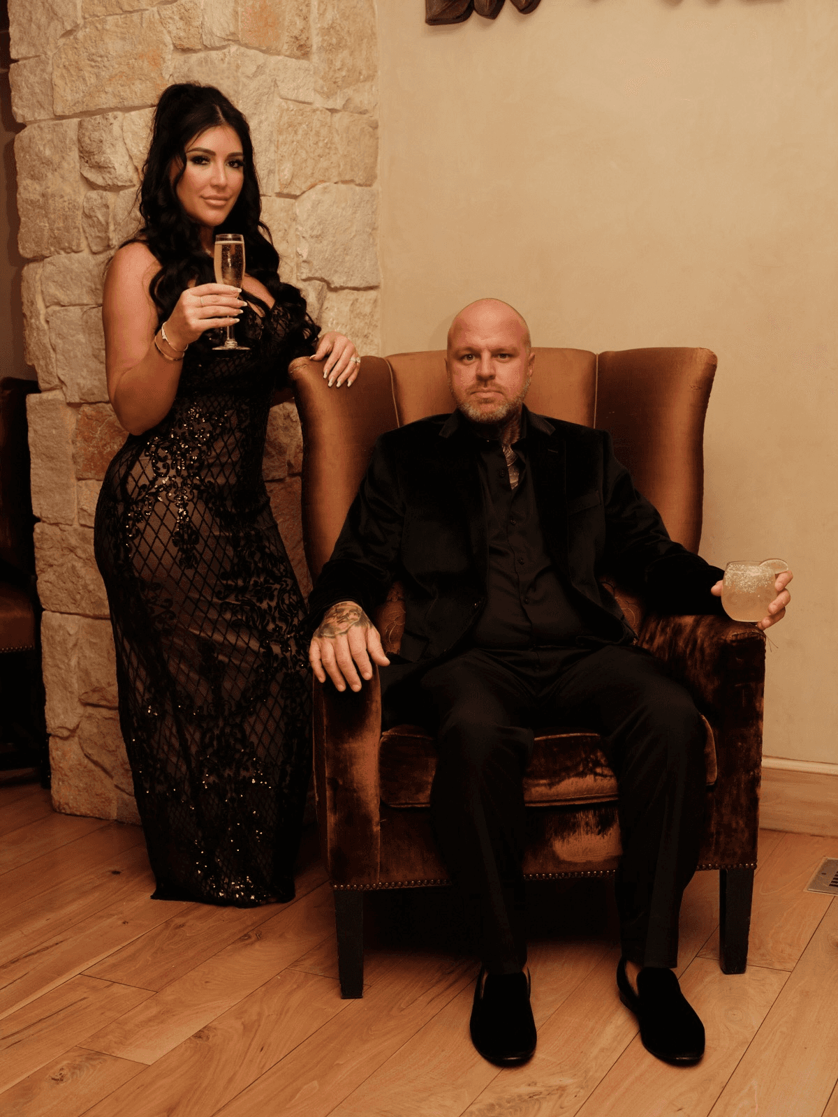 Elegant couple in formal attire holding drinks beside a stone fireplace in a luxurious indoor setting.