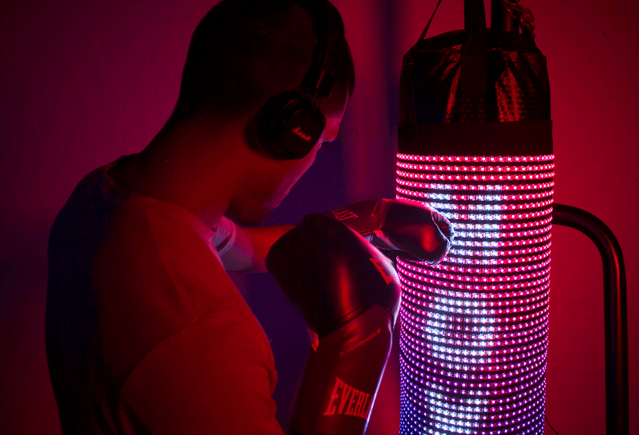 A person wearing black boxing gloves and over-ear headphones is positioned in front of a red LED boxing punching bag. The lighting in the scene is predominantly red, casting a glow on the bag and the person’s gloves. The person’s back is to the camera, and their head is slightly turned. The background is a solid dark red color.