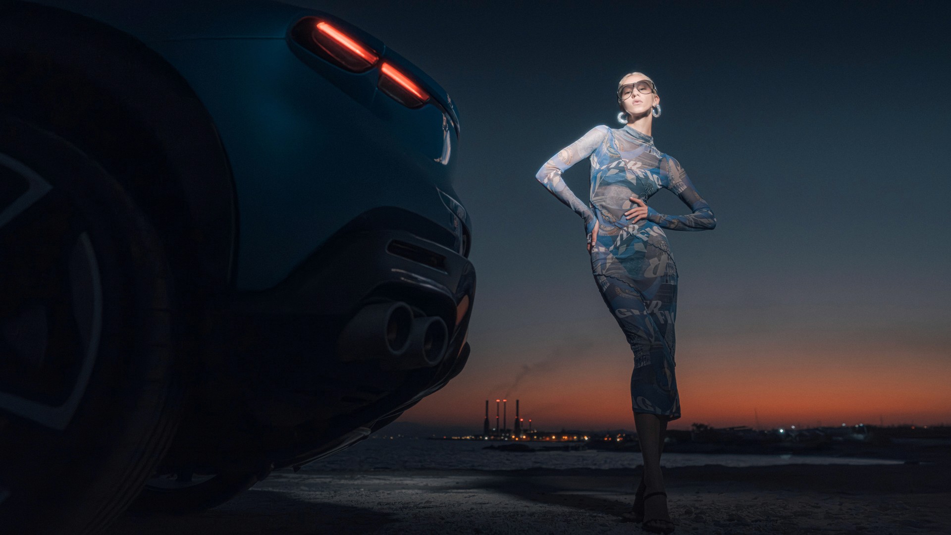 A model in a sheer printed dress and sunglasses poses with hand on hip beside the rear of a teal Ferrari Purosangue at dusk, low camera angle, with a coastline and distant industrial chimneys silhouetted against an orange-and-blue sunset.