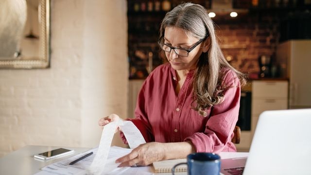 Mulher de cabelos grisalhos e óculos observa nota fiscal impressa, com notebook e papéis sobre a mesa.