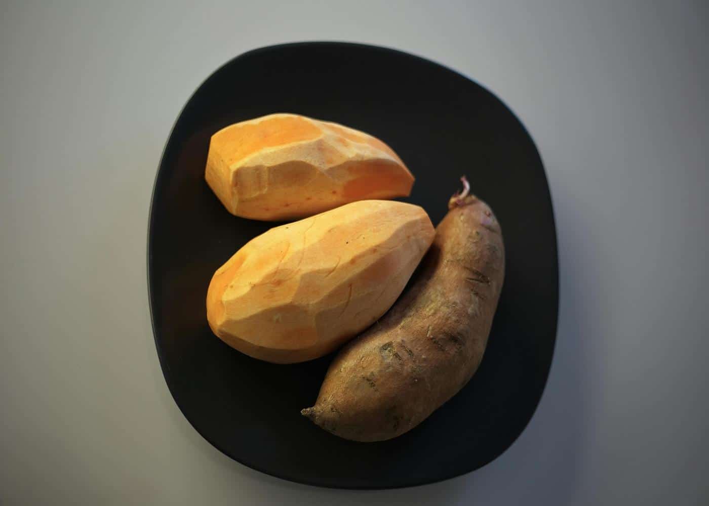 2 peeled raw sweet potatoes and 1 unpeeled sweet potato sitting on a black plate to showcase an alkaline diet