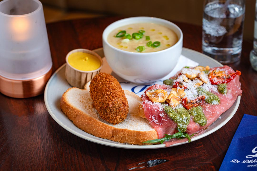 Een lunchgerecht met onder andere een brood waar een kroket op licht, een soepje en zalm