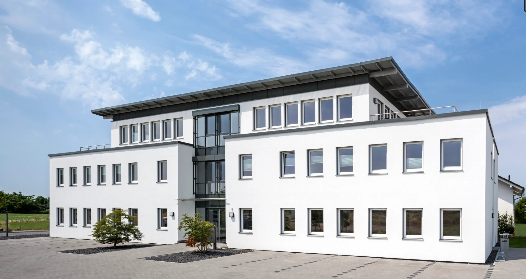 Modernes weißes Bürogebäude mit vielen Fenstern und einem Flachdach, an einem sonnigen Tag mit blauem Himmel und Wolken.