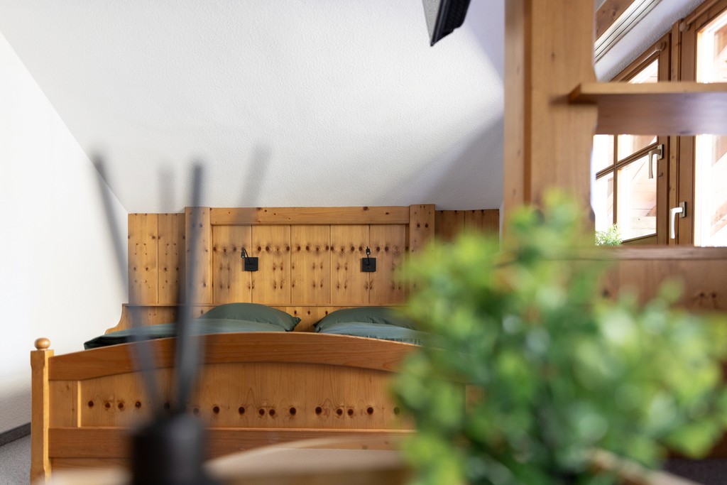 Ein gemütliches Schlafzimmer im Dachgeschoss verfügt über ein Holzbett mit grünen Kissen, sanftes Tageslicht, das durch ein kleines Fenster hereinströmt, und eine verschwommene Pflanze im Vordergrund, die einen Hauch von Grün verleiht.