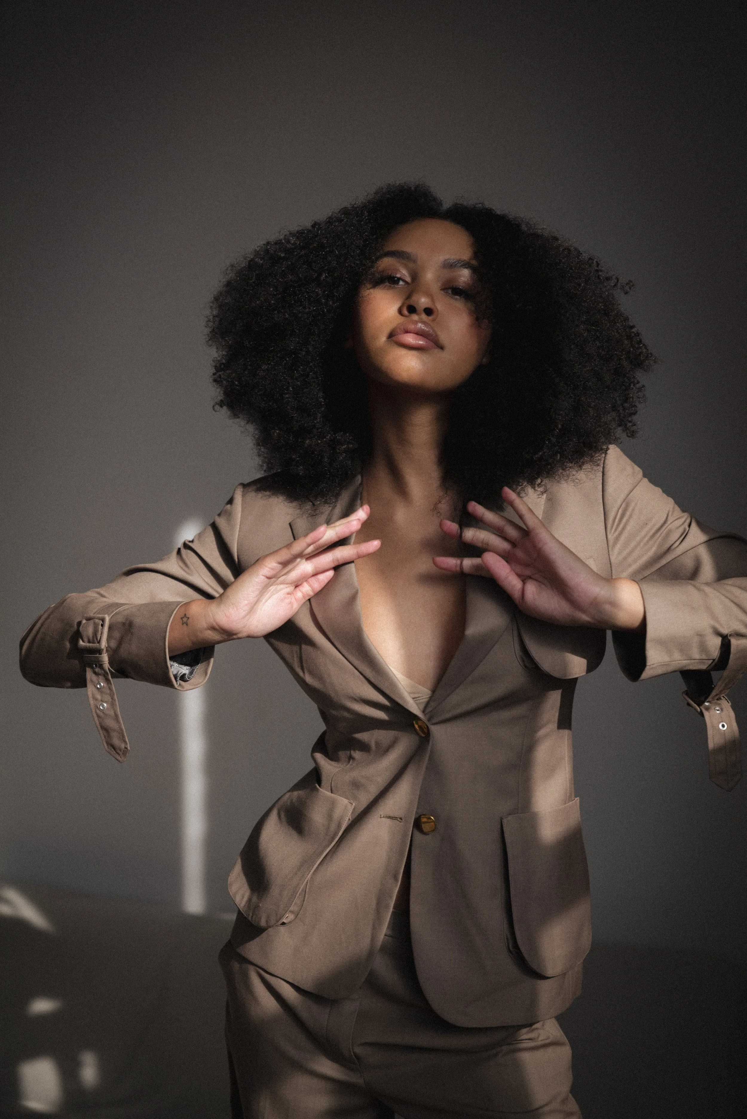 Woman with a voluminous afro in a tan suit, hands raised, dramatic studio light against a grey backdrop.
