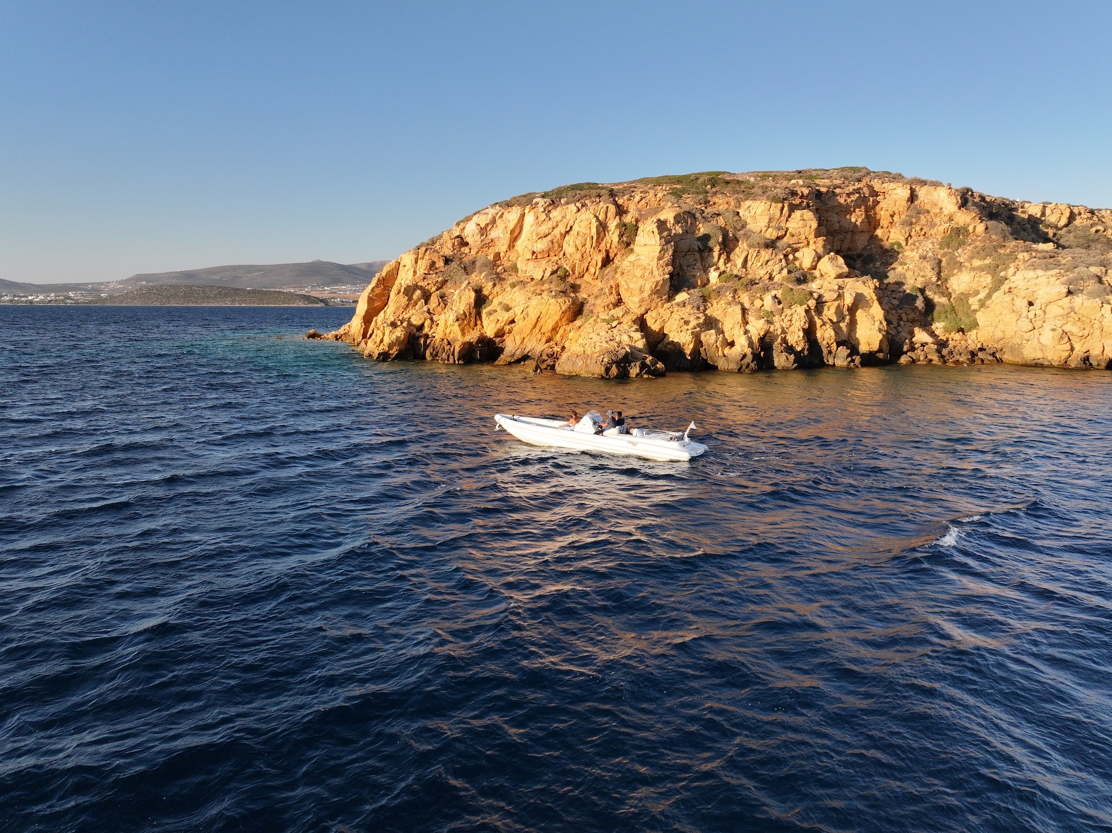 White speedboat cruising past dramatic golden cliffs in the Cyclades during golden hour with calm blue waters.