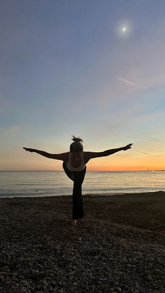 mujer con brazos abiertos y pierna levantada realizando postura de yoga frente al mar