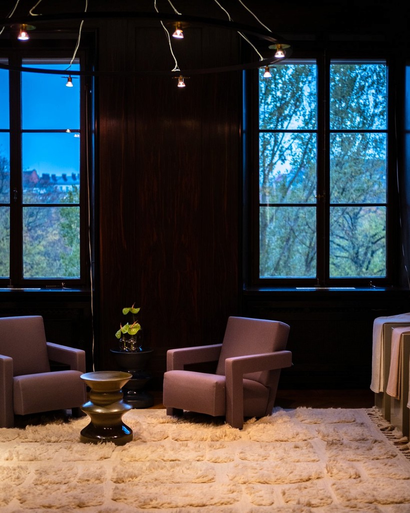 A cozy living room scene with two tan armchairs, a small black side table with a plant, a round decorative stool, and large windows showing trees outside during dusk.