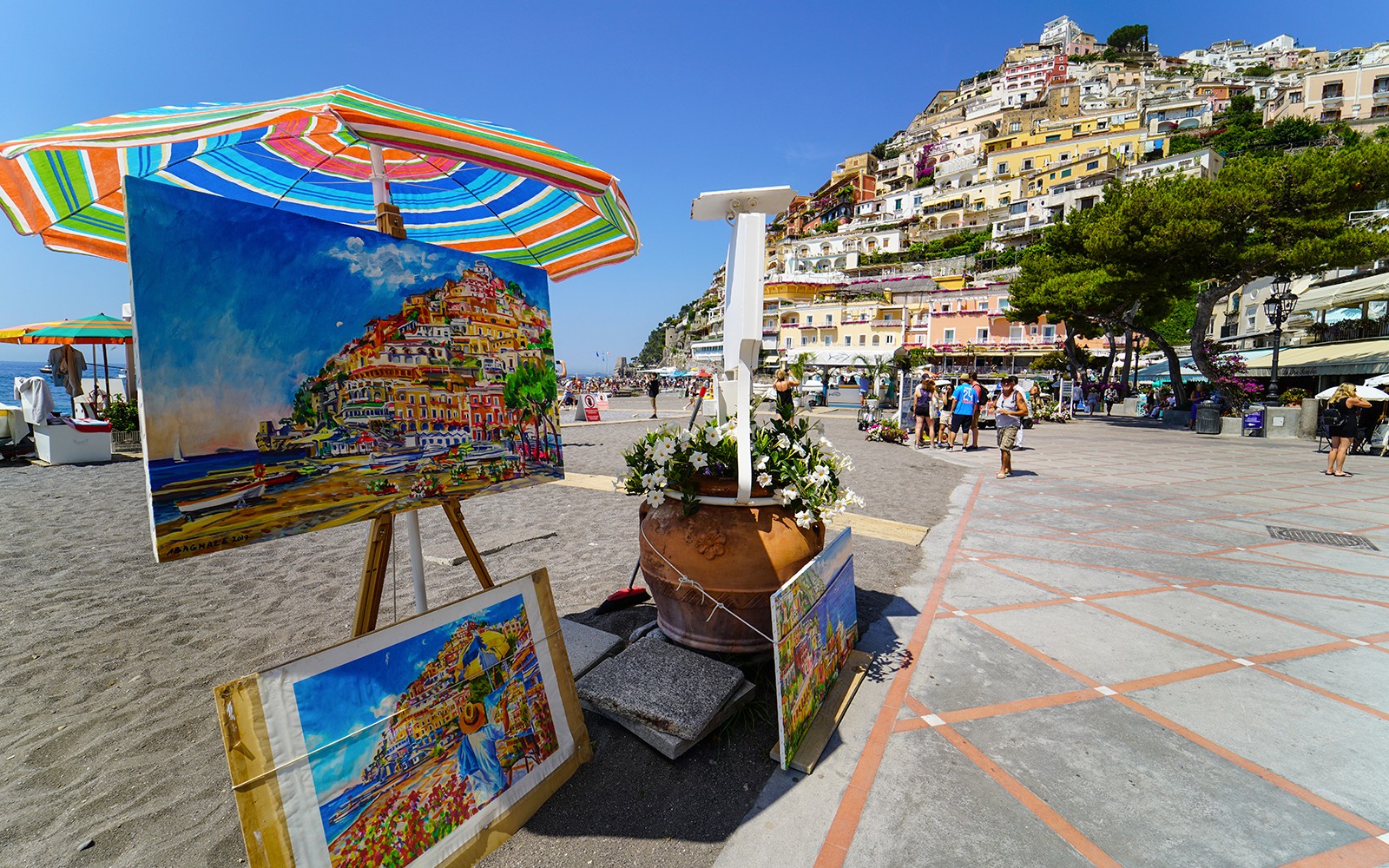 Färgglada målningar av Positano som visas på en strandpromenad, med hus på sluttningarna i bakgrunden.
