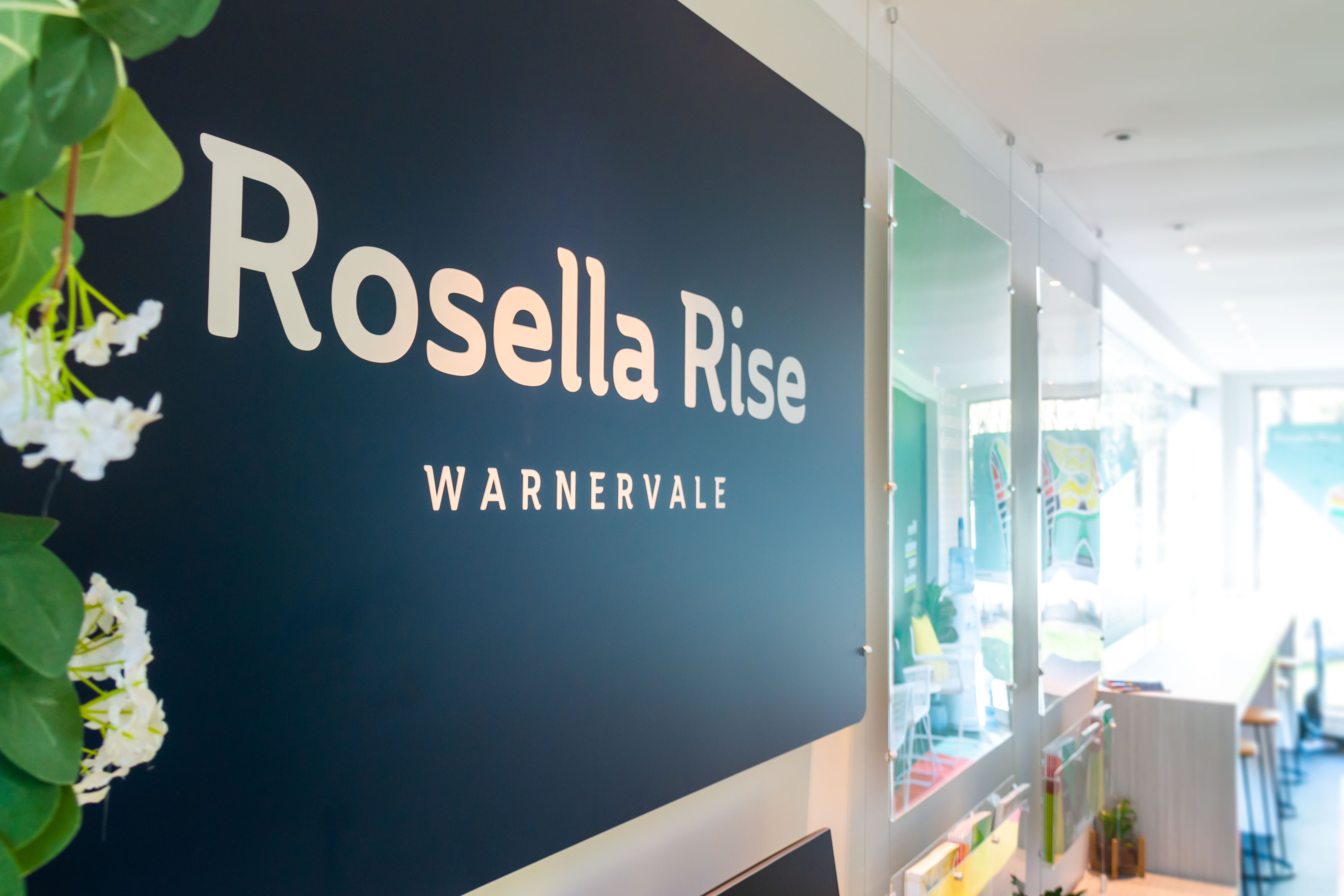 Blue sign reading "Rosella Rise Warnervale" with white text. Nearby are green plants and a bright, modern room with natural light.