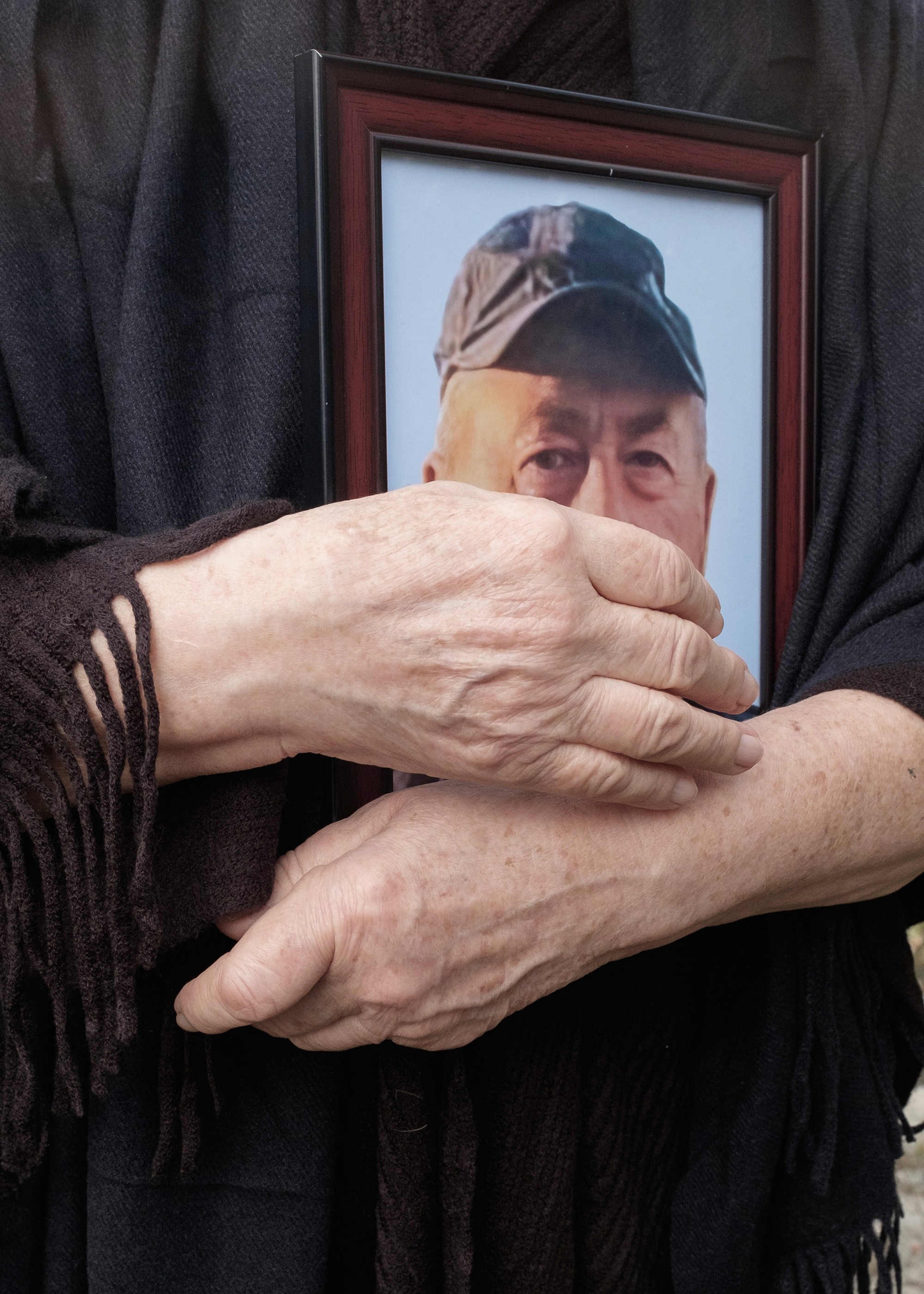 A mourner in black holds a framed portrait of Tamaz Ginturi, showing only his eyes and cap. Her hands are clasped tightly around the frame, capturing a moment of quiet grief