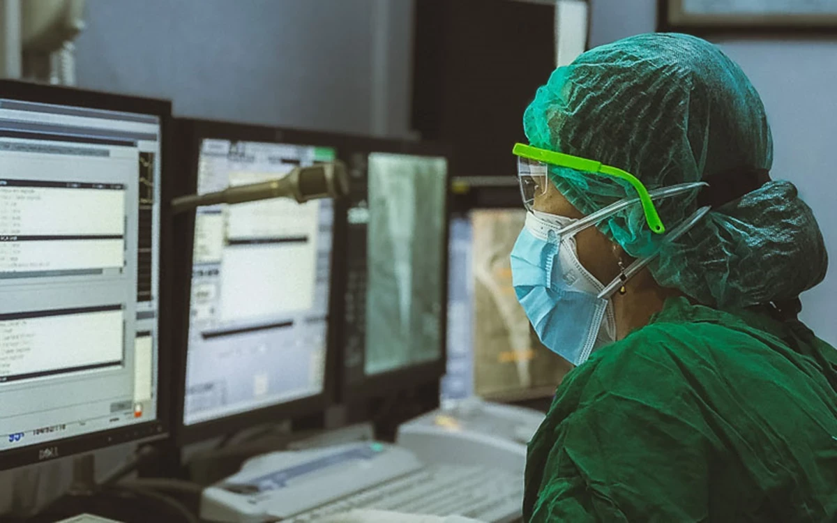 Medical professional wearing a green surgical cap, mask, and protective eyewear, working at multiple computer monitors in a clinical setting.