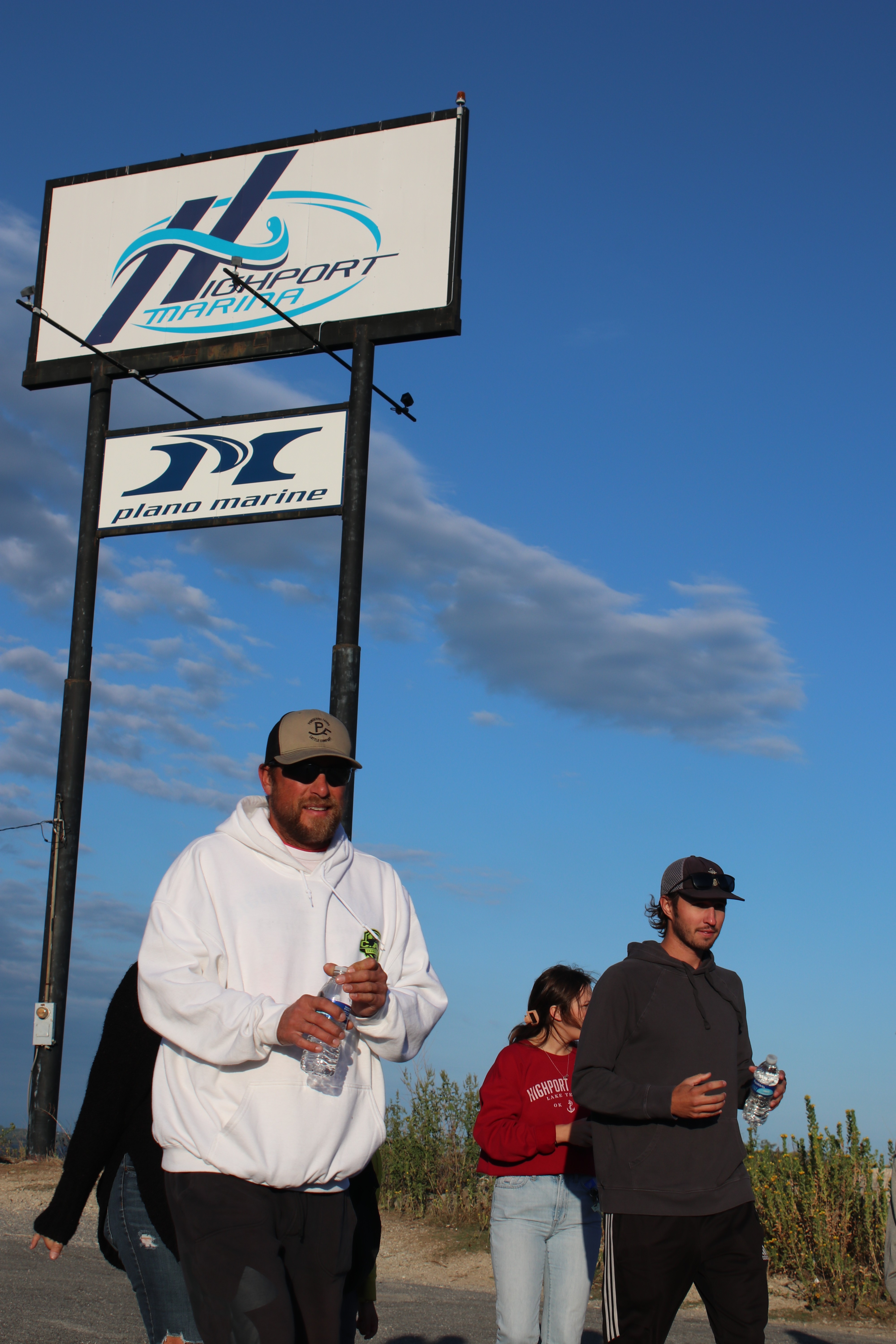 A group of people dressed in casual clothing, including hoodies and caps, walking along a roadside near a large sign reading "Plano Marine," with a clear blue sky and sparse clouds in the background, suggesting a sunny day.