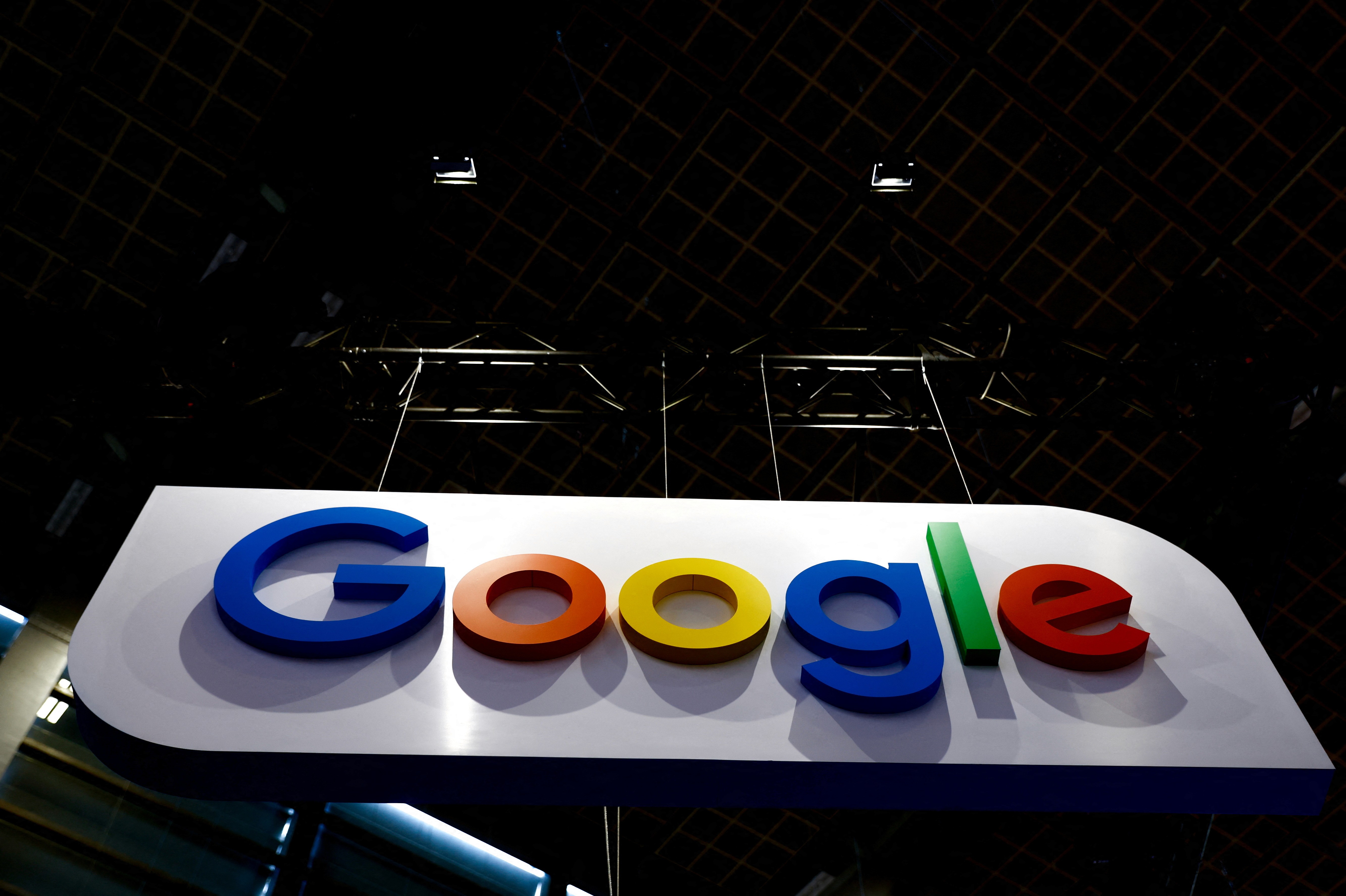 Google logo with bold, vibrant colors (blue, red, yellow, green) on a white sign, suspended from a ceiling structure in a dimly lit area.