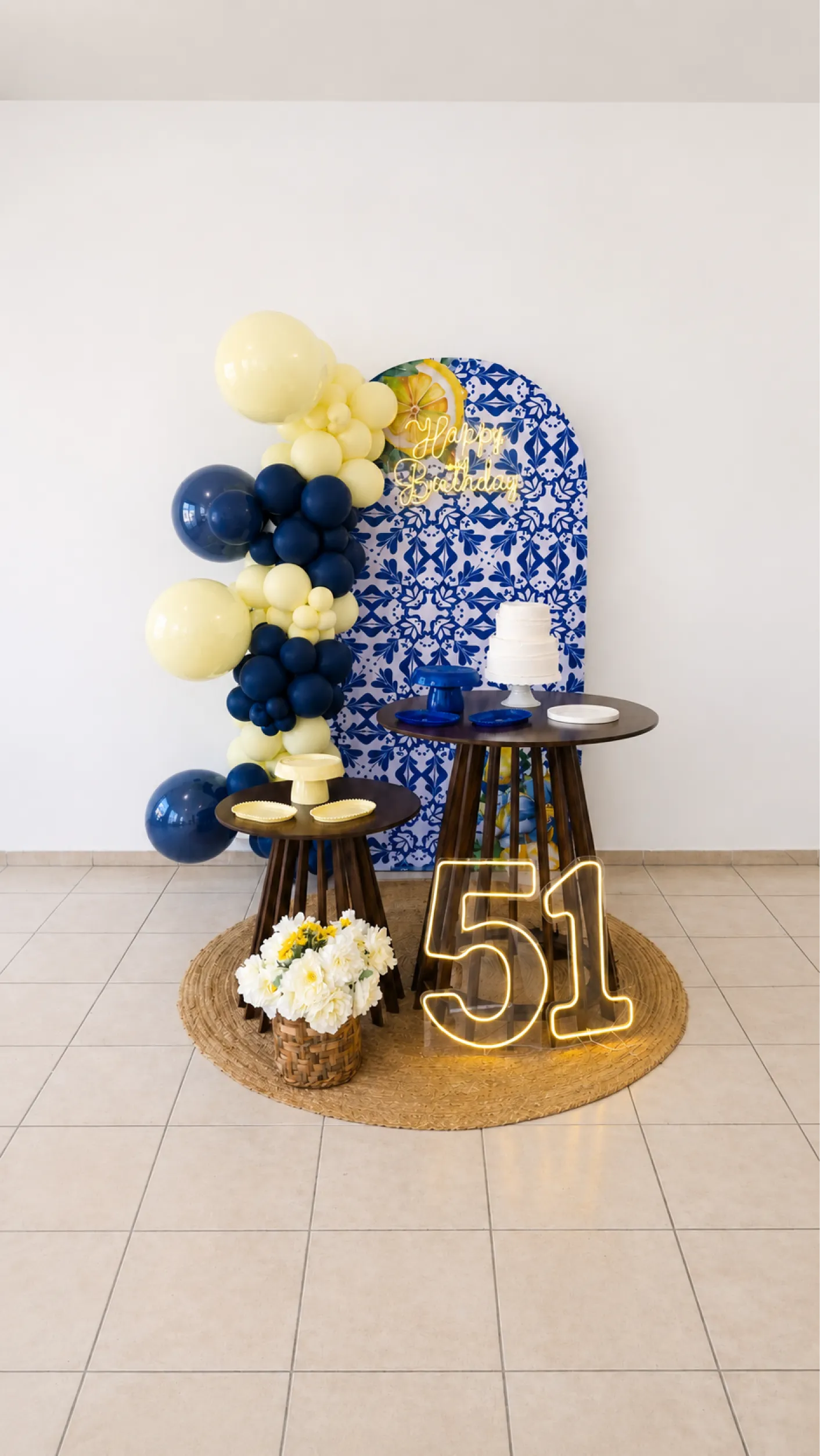 Decoração de aniversário com painel azul estampado, balões amarelos e azul-marinho, mesas escuras, flores brancas e número iluminado.